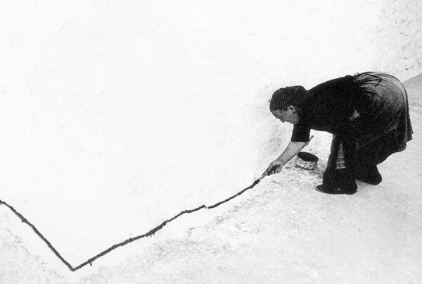 Imagen en blanco y negro de una mujer de los años sesenta rural, pintando una línea negra sobre el bajo de la fachada de una casa.