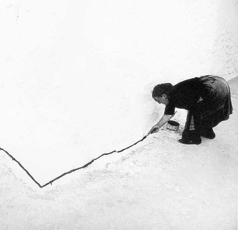 Imagen en blanco y negro de una mujer de los años sesenta rural, pintando una línea negra sobre el bajo de la fachada de una casa.