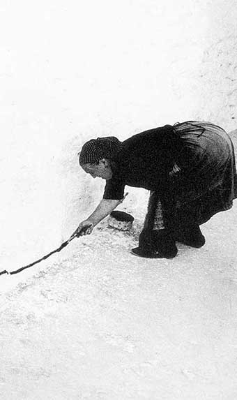 Imagen en blanco y negro de una mujer de los años sesenta rural, pintando una línea negra sobre el bajo de la fachada de una casa.