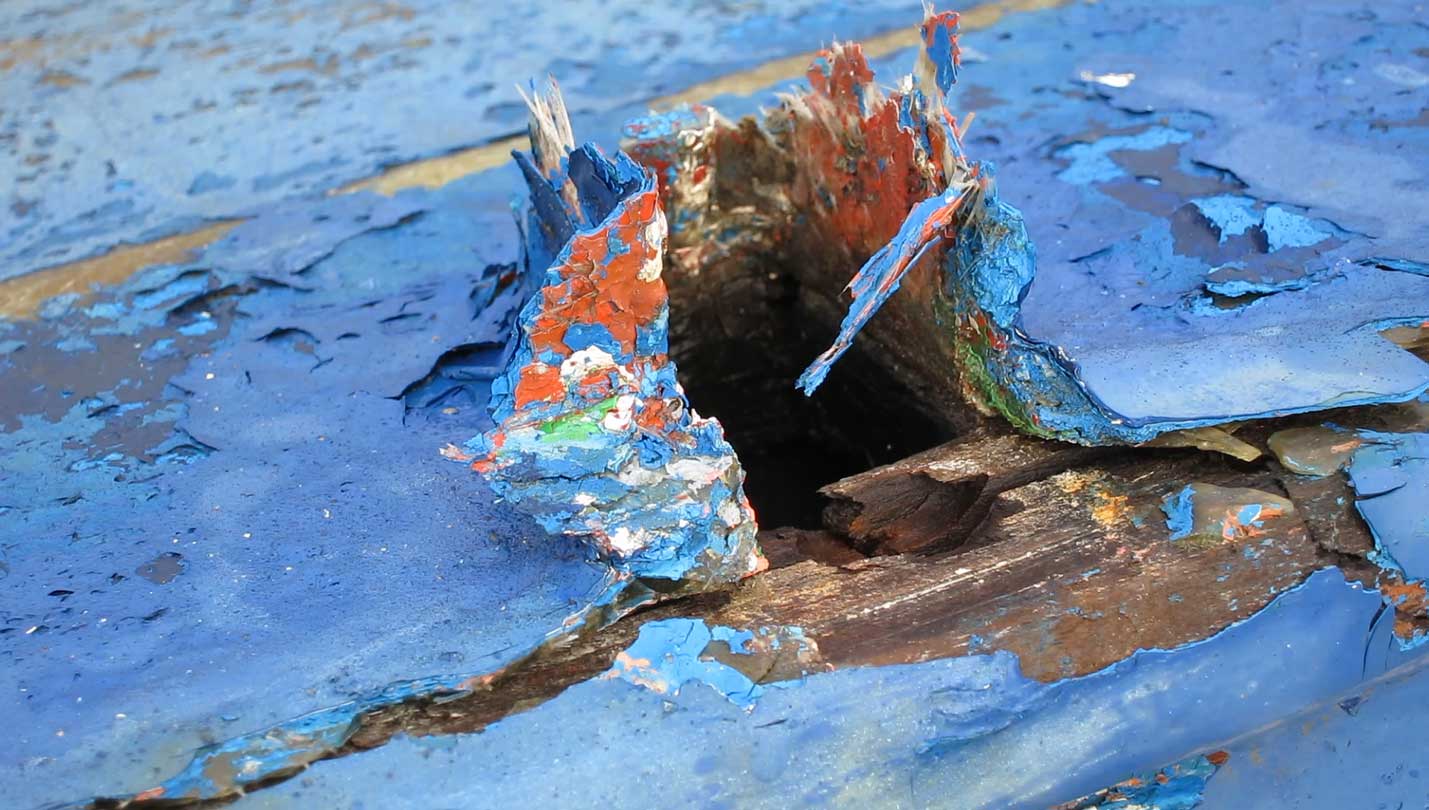 Detalle de una parte de una embarcación antigua deteriorada con el tiempo donde la pintura esta levantada y desgastada haciendo un juego de color con las anteriores capas de esta. Predomina el azul sobre rojo y verde de las capas anteriores.
