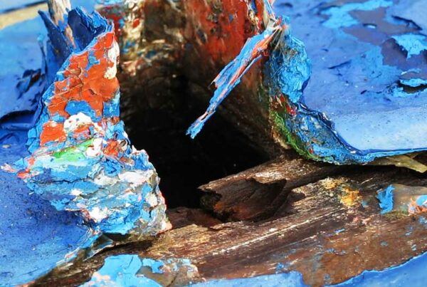 Detalle de una parte de una embarcación antigua deteriorada con el tiempo donde la pintura esta levantada y desgastada haciendo un juego de color con las anteriores capas de esta. Predomina el azul sobre rojo y verde de las capas anteriores.