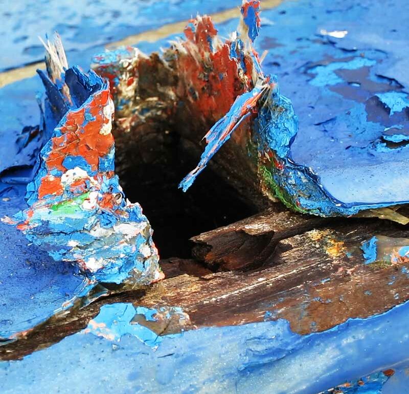 Detalle de una parte de una embarcación antigua deteriorada con el tiempo donde la pintura esta levantada y desgastada haciendo un juego de color con las anteriores capas de esta. Predomina el azul sobre rojo y verde de las capas anteriores.