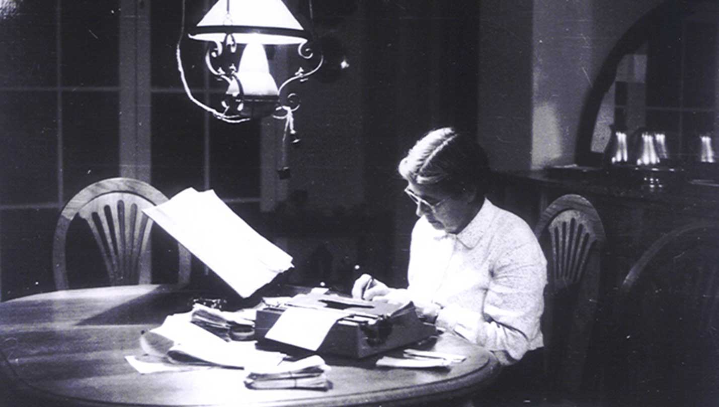 María Moliner sentada a la mesa en el comedor de su casa bajo una lámpara escribiendo con una máquina de escribir.